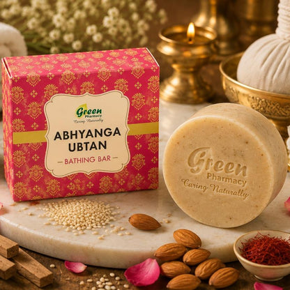 A bar of Green Pharmacy Abhyanga Ubtan bath soap displayed next to its pink patterned box on a marble slab, surrounded by bath ingredients like almonds, rose petals, cinnamon, and a cotton ball in a spa-style setting.