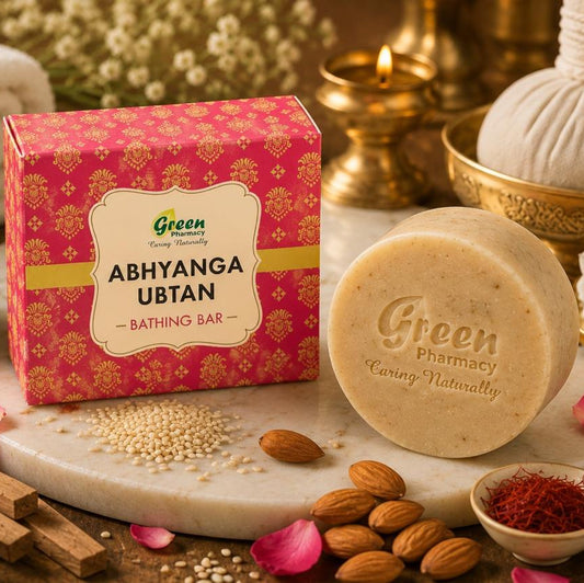 A bar of Green Pharmacy Abhyanga Ubtan bath soap displayed next to its pink patterned box on a marble slab, surrounded by bath ingredients like almonds, rose petals, cinnamon, and a cotton ball in a spa-style setting.