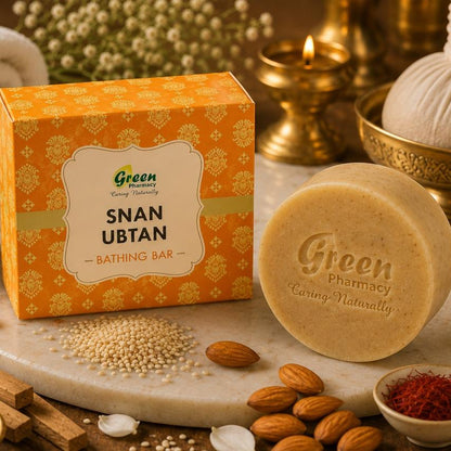 A bath bar product display featuring a round beige bath bar labeled 'Green Pharmacy Snan Ubtan' next to its orange patterned box, styled with almonds, spices, flowers, and traditional bath accessories on a marble surface.